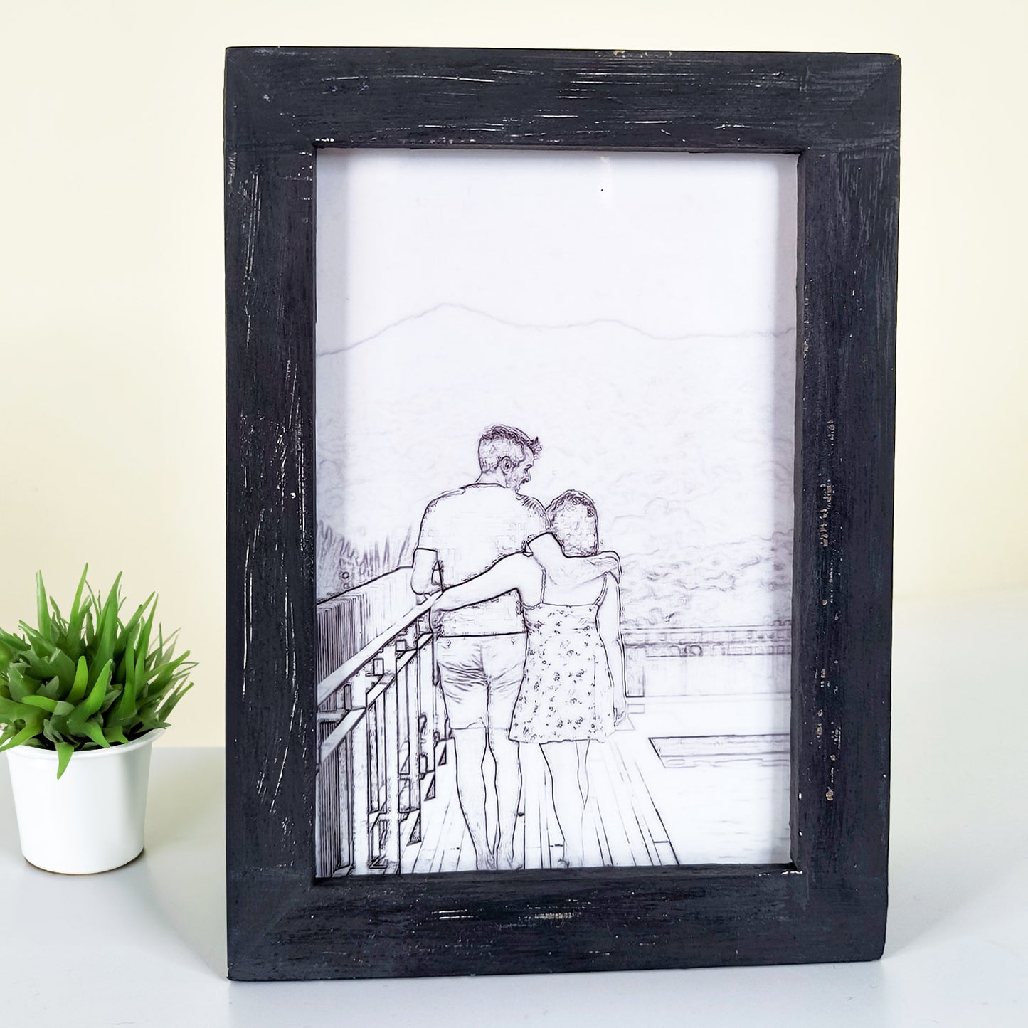 A framed pencil sketch of a couple standing on a balcony, with the man’s arm around the woman’s shoulder. A small potted plant is placed beside the frame.