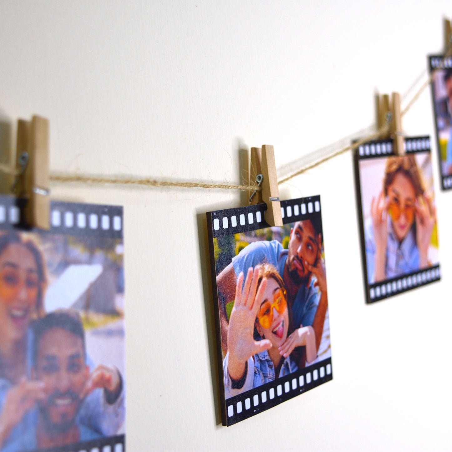 A string of reel photo wall decor is displayed, each film-styled print clipped to the string with a small wooden clothespin against a white wall. The photos show a happy pair of friends making playful gestures and smiling brightly.