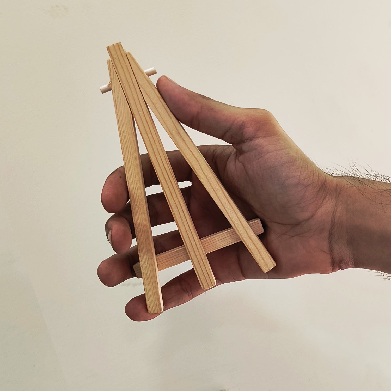 A person's hand is holding a small wooden structure resembling a simple, triangular ladder. The background is plain and light-colored, keeping the focus on the object and the hand.