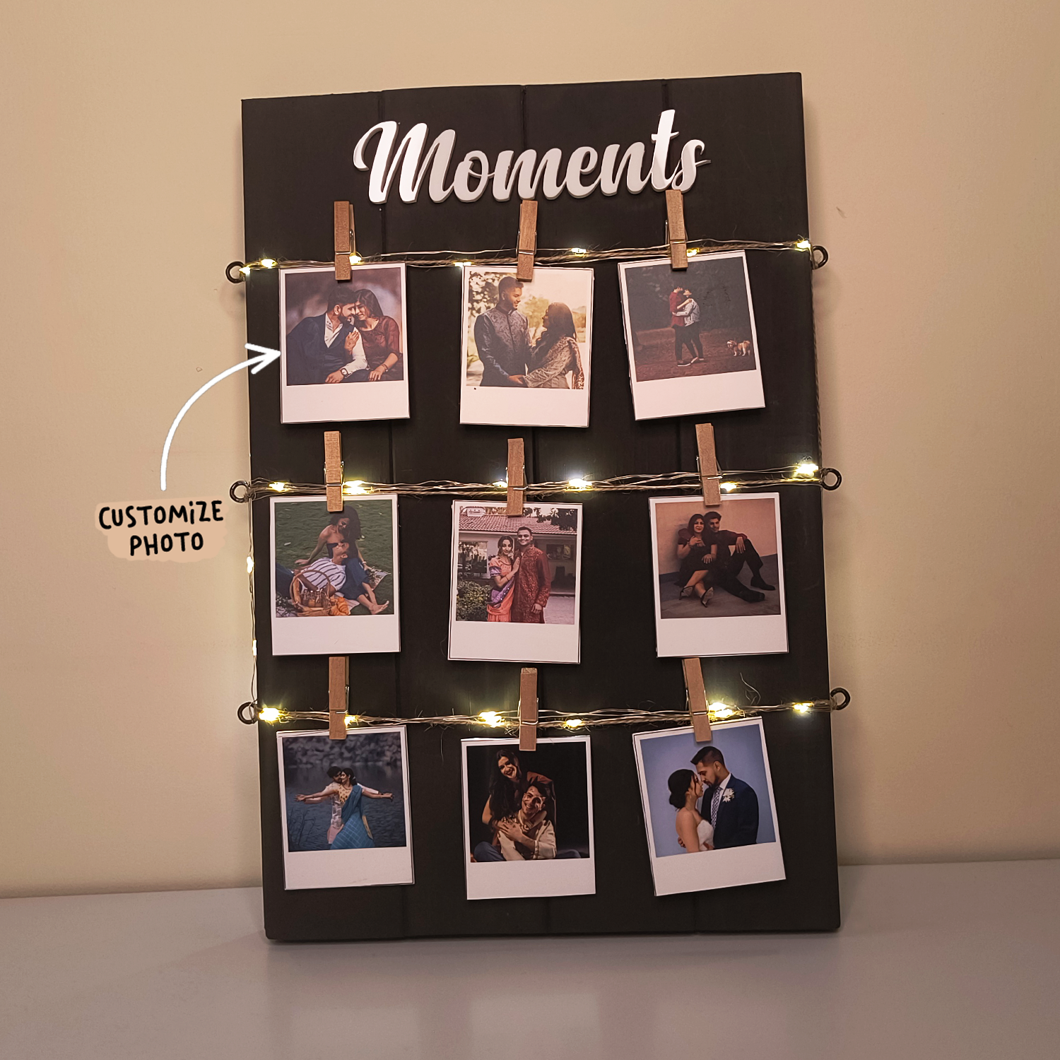 A photo display board titled "Moments" features nine polaroid-style photos clipped to strings of lights with wooden clothespins. An arrow pointing to the top-left photo has text that says "Customize Photo." The board rests against a beige wall on a flat surface.