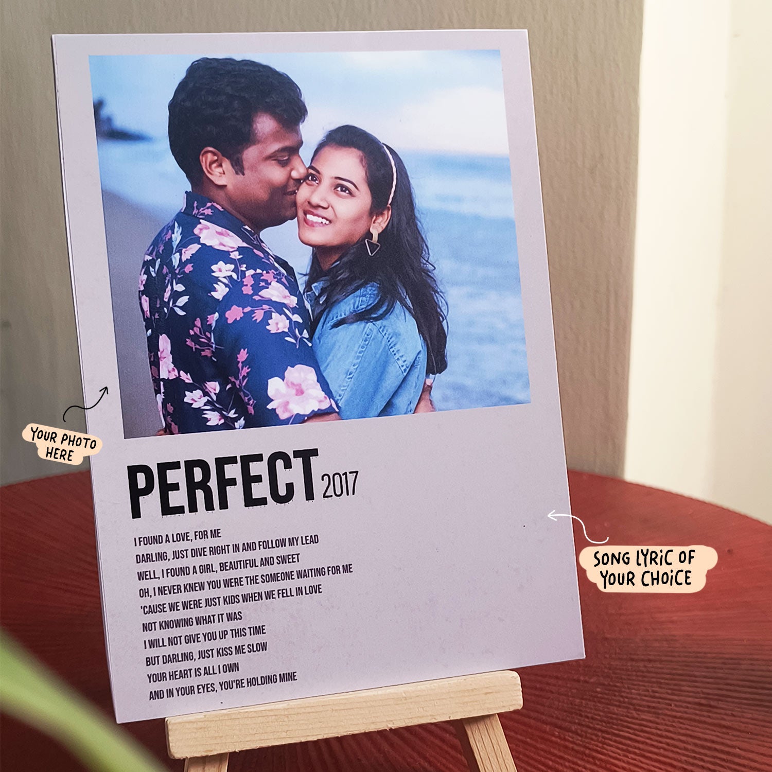 A Song Lyrics Poster with framed photo of a couple standing on the beach is placed on a wooden surface. The man is wearing a floral shirt, and the woman is wearing a white sweater, both smiling. The frame reads "PERFECT 2017" with customizable spots for a photo and a song lyric.