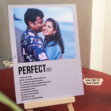 A Song Lyrics Poster with framed photo of a couple standing on the beach is placed on a wooden surface. The man is wearing a floral shirt, and the woman is wearing a white sweater, both smiling. The frame reads 