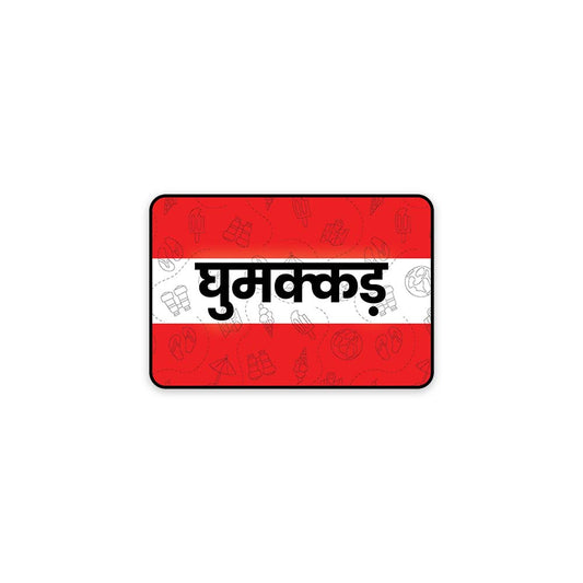 A rectangular sticker with a red background and a white horizontal stripe in the middle. In the center of the white stripe, black Devanagari text reads "घुमक्कड़" which translates to "Wanderer." The red background has faint travel-themed illustrations.